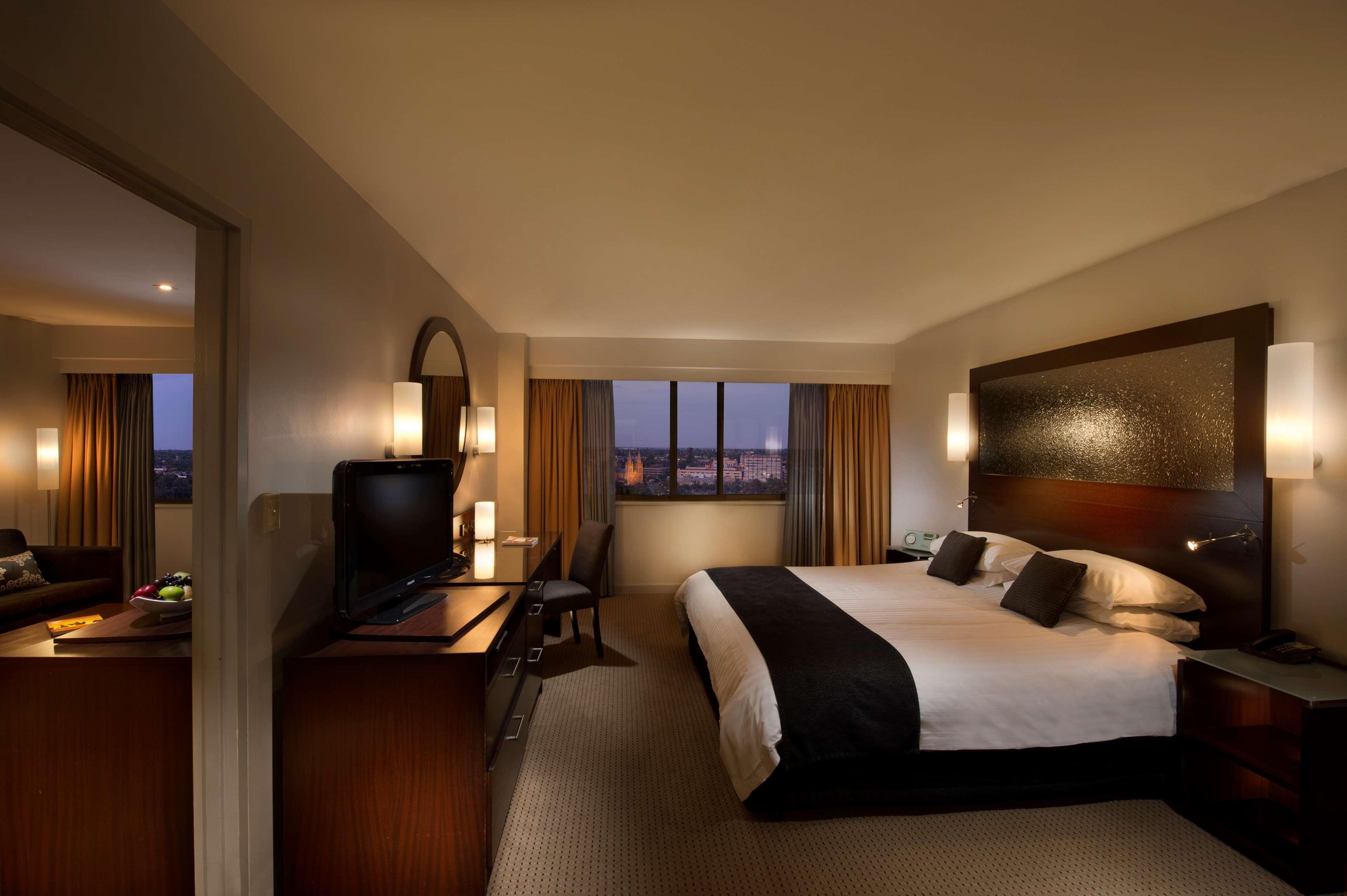 מלון Stamford Plaza אדלייד מראה חיצוני תמונה The photo features a modern hotel room that includes a large bed with dark and light bedding, complemented by two pillows. The room is well-lit with warm lighting and has a contemporary aesthetic. To one side, there is a wooden dresser with a flat-sc