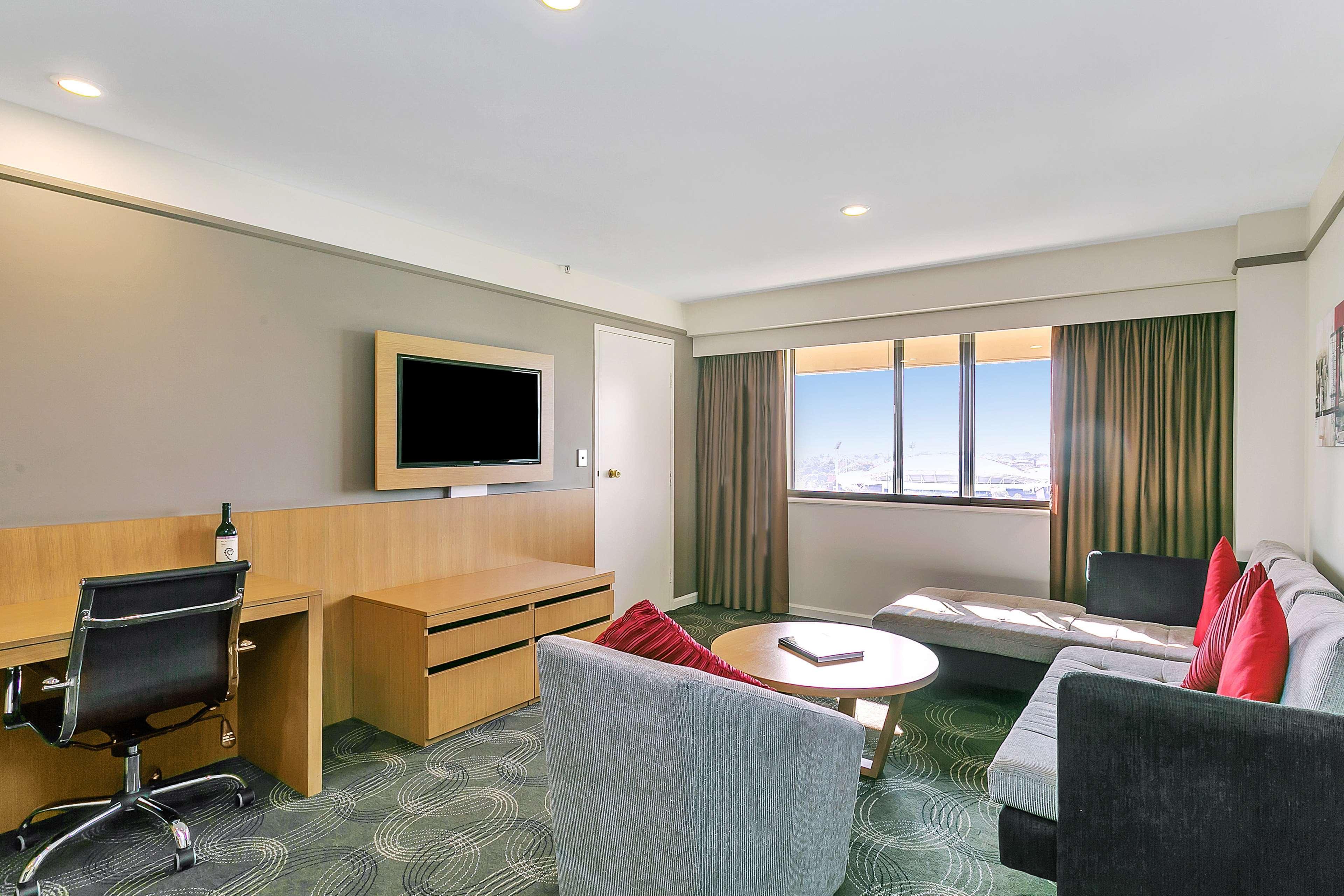 מלון Stamford Plaza אדלייד מראה חיצוני תמונה The photo shows a modern hotel room or suite that features a cozy seating area. There is a comfortable sofa with red accent pillows, a round coffee table in the center, and a sleek desk with an office chair. On the wall, there is a flat-screen TV mou
