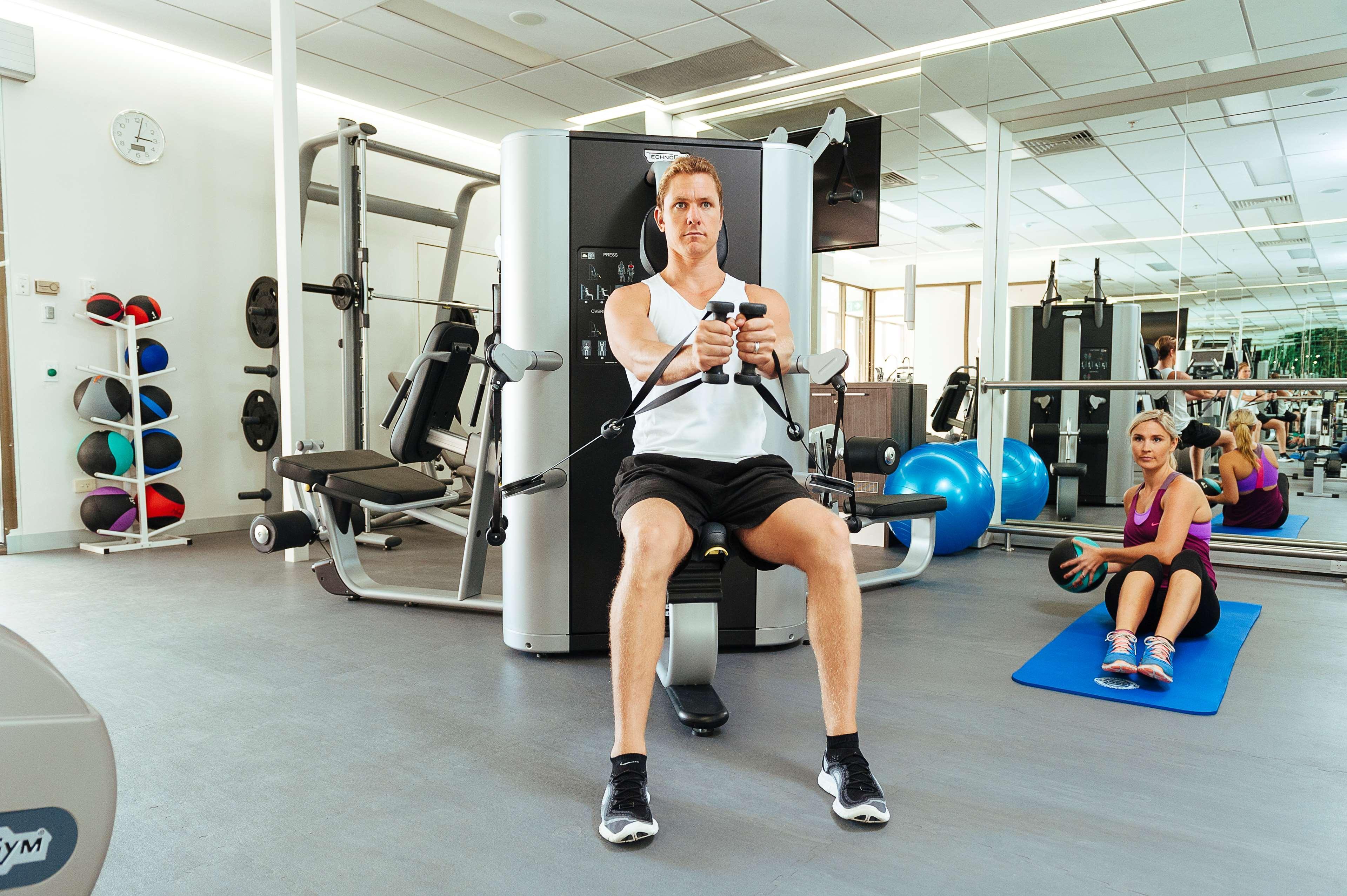 מלון Stamford Plaza אדלייד מראה חיצוני תמונה The photo shows a gym setting with two individuals engaged in workout activities. 

In the foreground, a man is seated on a machine, using the handles to perform a rowing exercise. He is wearing a sleeveless shirt and shorts, indicating an active wor