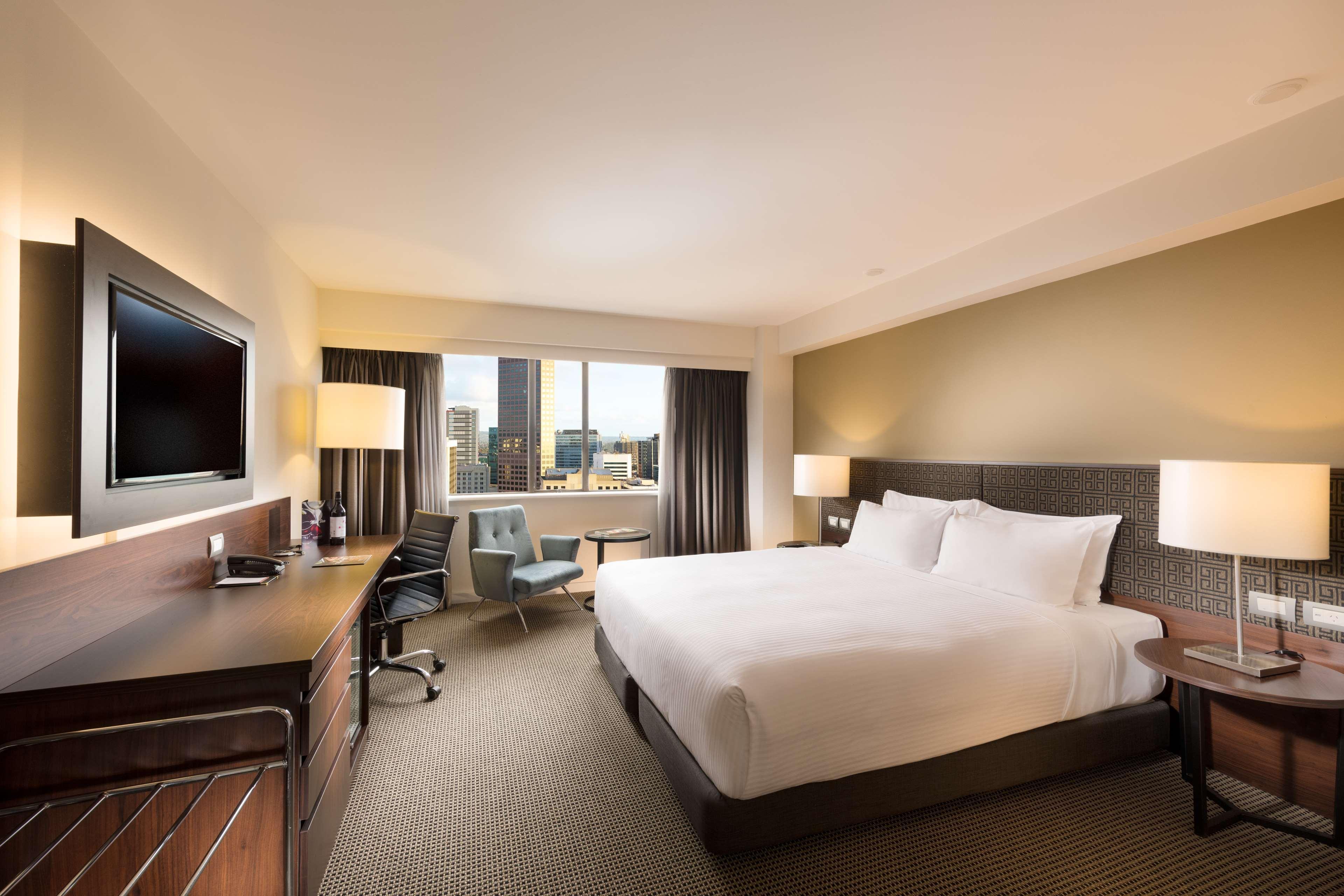 מלון Stamford Plaza אדלייד מראה חיצוני תמונה The photo shows a modern hotel room with a contemporary design. It features a large bed with white linens, a bedside table on either side with lamps, and a dark wood accent wall. There's a work desk with an office chair and a flat-screen television m