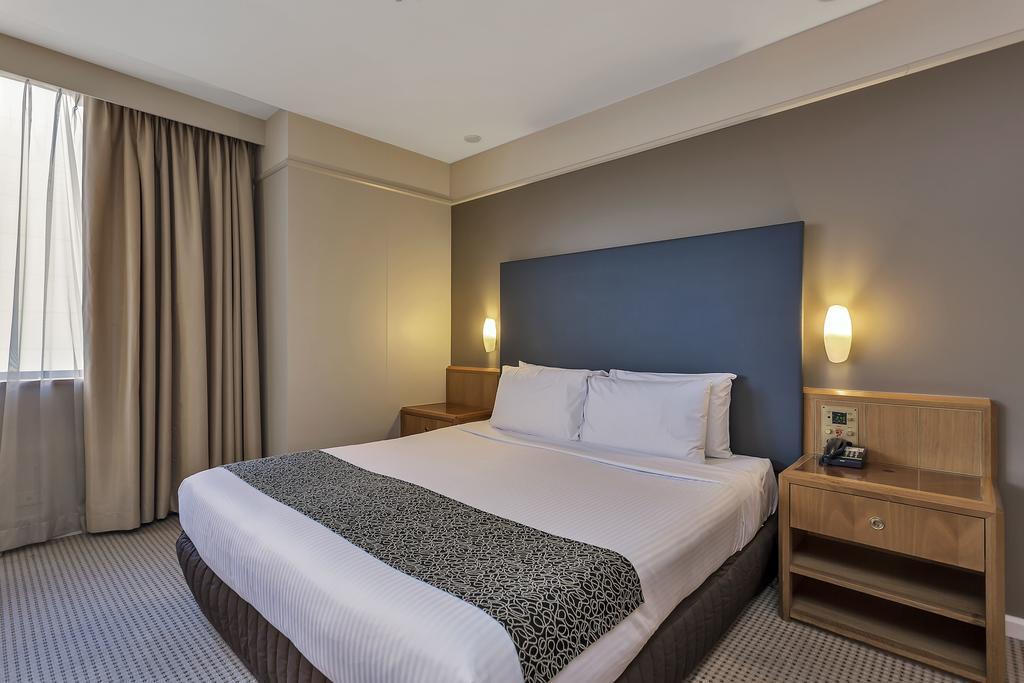 מלון Stamford Plaza אדלייד מראה חיצוני תמונה The photo shows a modern hotel room. It features a double bed with a white duvet and a decorative grey and black throw. The headboard is a solid dark color. There are two bedside tables on either side of the bed, each with a lamp. Natural light enter
